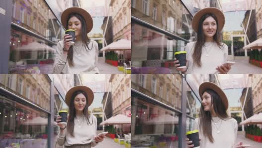 迷人的肖像迷人的快乐的年轻女子戴着时髦的帽子，走在现代建筑与街头咖啡馆之间，一只手拿着咖啡和电话在另一个高清在线视频素材下载