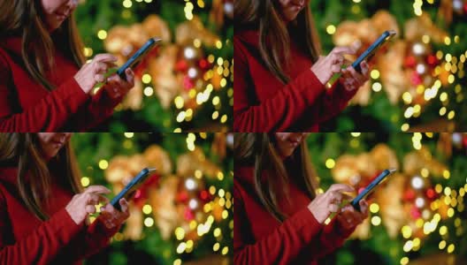 穿着红色毛衣在圣诞树前玩手机的成熟女人高清在线视频素材下载