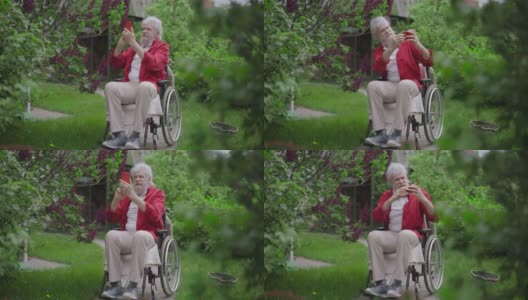 开朗的残疾老人在春夏季花园与盛开的大树自拍。快乐正面白种人老人的肖像享受大自然的户外。缓慢的运动。高清在线视频素材下载