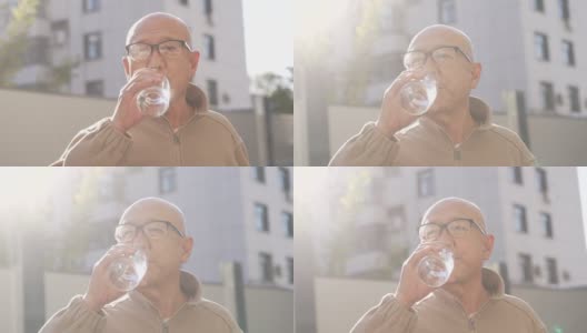 一位亚洲老人，戴着老花镜，站在外面的阳光下喝水高清在线视频素材下载