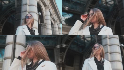 一个戴着眼镜、穿着白色外套的女孩站在市中心一家酒店的背景下，调整着自己的眼镜高清在线视频素材下载