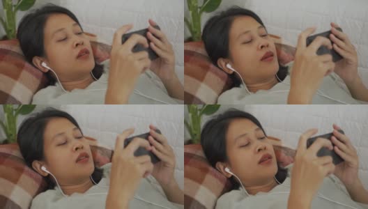 亚洲女人躺着，戴着耳机，用智能手机玩游戏高清在线视频素材下载