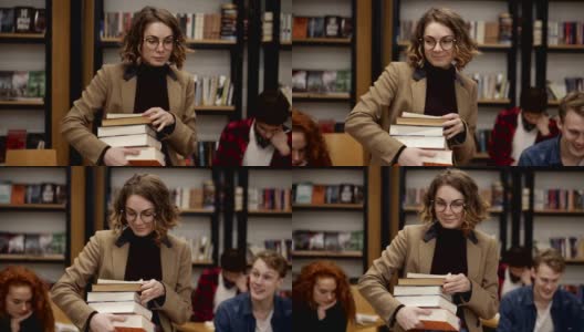 一个穿着棕色夹克，戴着眼镜的女孩拿着书架上的一堆书走在路上的慢镜头。其他同学坐着，在学院、大学图书馆里学习。前视图高清在线视频素材下载