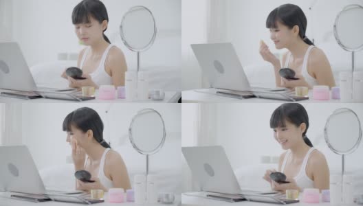 美丽的年轻亚洲女人学习化妆与粉扑在脸颊上看笔记本电脑与教程在线，女性教学和解释化妆与化妆品流。高清在线视频素材下载