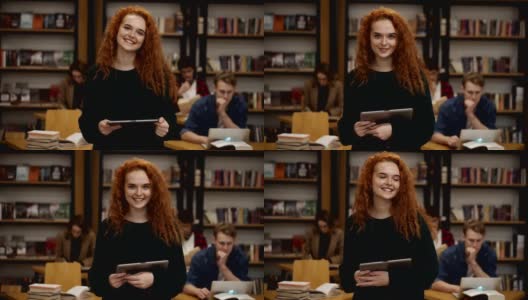 这是一幅迷人的长红头发欧洲女学生的肖像，她站在高中图书馆里，微笑着看着相机，手里拿着平板电脑。教育、文学和人的观念。背景模糊的同学高清在线视频素材下载