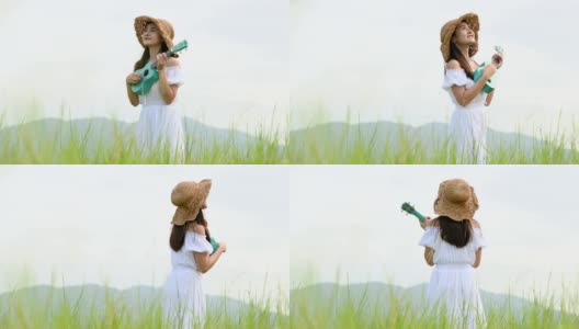 亚洲快乐的女人在白色的衣服弹奏尤克里里吉他在绿色的草地与山的背景在夏天。音乐家美女放松与乐器。乡村的生活方式高清在线视频素材下载