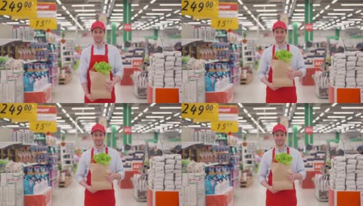 穿着红色制服的售货员拿着装着新鲜蔬菜的牛皮纸包站在超市里。高清在线视频素材下载