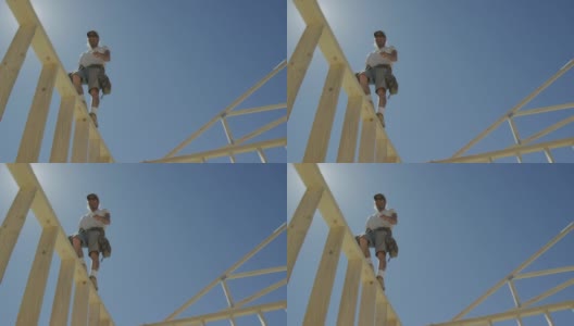 在一个晴朗、阳光明媚的日子里，一个40多岁的白人男性建筑工人在一个框架房屋的顶部行走，身后是一台液压起重机高清在线视频素材下载