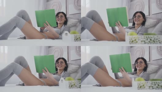 漂亮的小女孩戴着眼镜躺在卧室的床上，她微笑着读着一本有趣的书。穿着时髦的灰色衣服的女孩在早晨读一本有趣的书高清在线视频素材下载