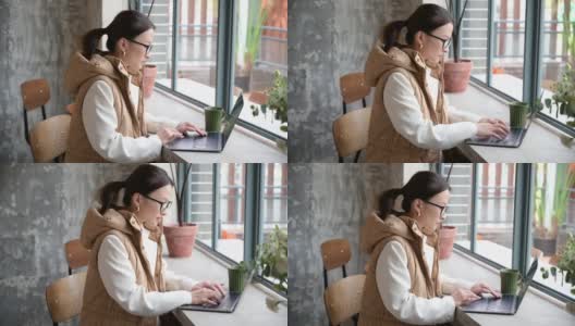 漂亮的亚洲华人女商人戴着眼镜，在咖啡馆靠窗的地方用笔记本电脑做自由职业者。休息时间上网工作。敲击键盘。年轻的学生在咖啡馆学习高清在线视频素材下载