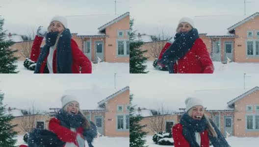 快乐的年轻微笑的女人扔雪球，并被一个击中。穿红外套的女孩和她的伴侣享受冬天。在缓慢的运动。田园小屋后院里的一天。高清在线视频素材下载