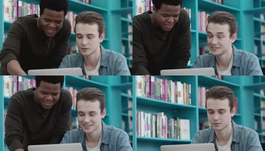 一个年轻的白人在大学图书馆用笔记本电脑做作业。他的非洲朋友和他一起帮忙完成任务高清在线视频素材下载