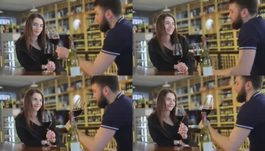 一个男人和一个女人正在用玻璃杯喝红酒，一个餐厅的内部，一个年轻的男人和一个女孩在一个餐厅的桌子上喝葡萄酒，背景是一个葡萄酒架子，一个男性嬉皮士高清在线视频素材下载