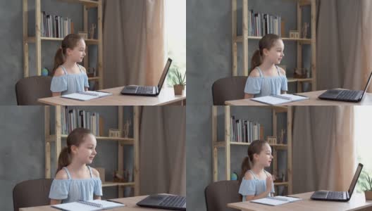 小白人女孩微笑和挥手，而坐在家里的桌子上与笔记本电脑。在线交流，远程学习。高清在线视频素材下载