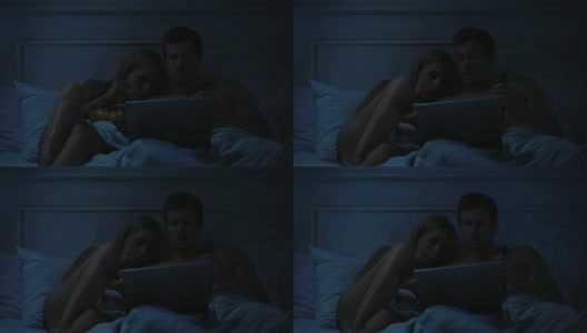 女士和男士坐在黑房间的床上，用手提电脑看恐怖电影高清在线视频素材下载