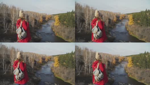 一名身穿红色雨衣、头戴米色帽子、肩上背着灰色背包的女子站在山顶眺望远方。风使头发变长。河流，绿色和金色的树叶的树木高清在线视频素材下载
