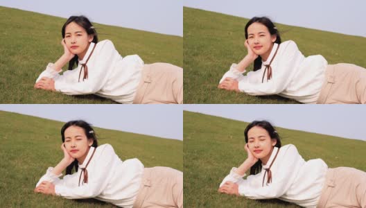 一名美丽的中国年轻女子躺在绿草如茵的草地上，手捧着头微笑着对着镜头，享受着春天的微风，4k镜头，慢镜头。高清在线视频素材下载