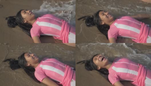 快乐的亚洲女孩穿着粉红色的泳衣躺在沙滩上溅起的水，慢动作。高清在线视频素材下载