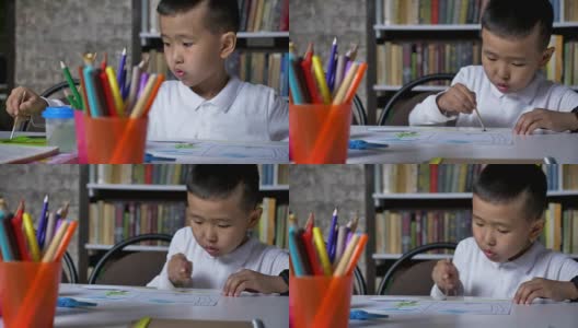 年轻的亚洲小男孩在纸上画画，坐在桌子后面，孩子做作业，书架的背景高清在线视频素材下载