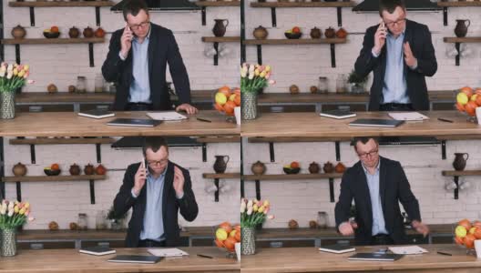 一位戴着眼镜、穿着西装、怒气冲冲的企业家站在厨房里一边打手机一边打着手势。他把手机扔在桌子上，把笔记本从桌子上推下来高清在线视频素材下载