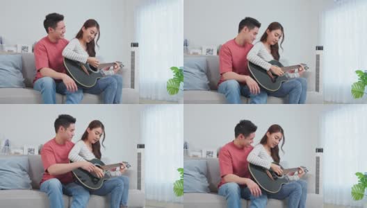 亚洲年轻情侣在家里的客厅里一起弹吉他。迷人浪漫的新婚，活跃浪漫的男人坐在沙发上教漂亮的女朋友在家里弹琴。高清在线视频素材下载