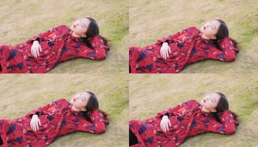 一名身穿红色连衣裙的中国年轻女子双手抱头躺在草地上的肖像，4k镜头，慢镜头。高清在线视频素材下载