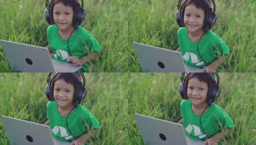 小女孩戴着耳机，在绿草地上用笔记本电脑社交媒体。乡村景色。在夏天的休闲时间高清在线视频素材下载