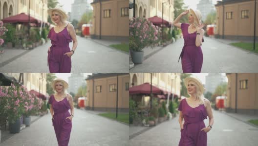 一个积极的女人穿着紫色的连衣裤，戴着草帽，慢动作地走在镇上的户外四处张望。前视图肖像自信优雅苗条美丽的成年女士漫步在城市的早晨高清在线视频素材下载