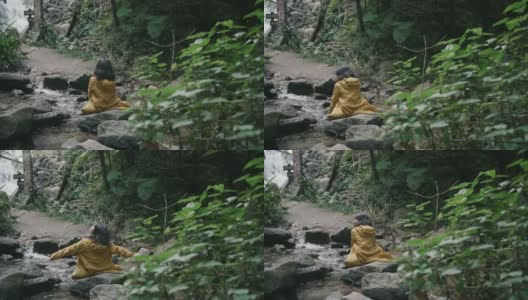 穿着黄色雨衣的亚洲女孩坐在瀑布，张开双臂。高清在线视频素材下载