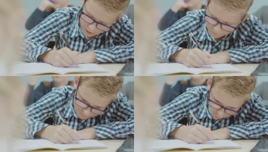 小学课堂:一个聪明的白人男孩戴着眼镜在练习本上写字。学习科学和创造性思维的多元聪明儿童群体高清在线视频素材下载