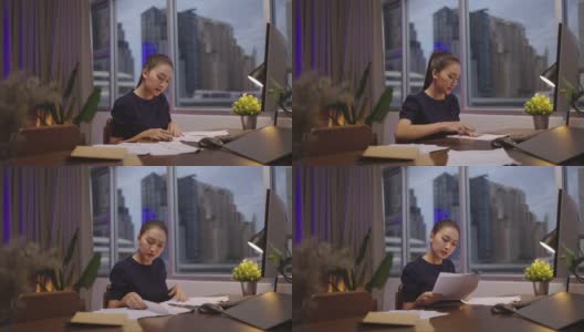 一名亚洲女性坐在她的办公桌前，在家里用电脑和文件工作到深夜。高清在线视频素材下载