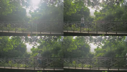 在温暖的晨光下，与身着运动服的亚洲中年女性在绿树成荫的自然公园桥上慢跑。高清在线视频素材下载