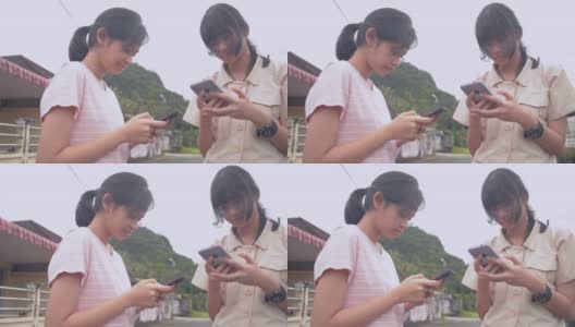 两个亚洲可爱的少女女友一起站在屋外，用智能手机发短信。都市的现代生活。社会网络交流。高清在线视频素材下载