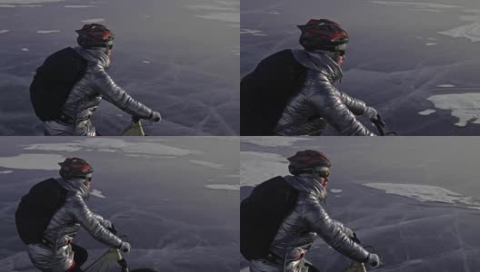 一个女人在冰上骑自行车。这个女孩穿着银色的羽绒服，背着自行车背包，戴着头盔。用四旋翼无人机拍摄。自行车的轮胎上布满了钉子。高清在线视频素材下载