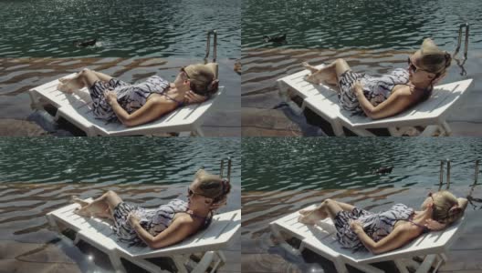 一个女人躺在太阳床上，戴着太阳镜，披着波西米亚风格的丝绸披肩。女孩休息在一个洪水木头水下码头。鸭子和天鹅在附近游泳。高清在线视频素材下载