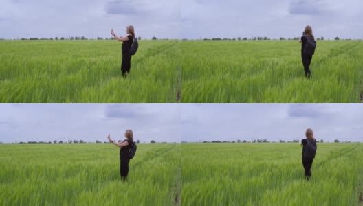 一名女游客在乡间徒步行走，站在一片生长着小麦的绿色田野上，用手机拍照。高清在线视频素材下载