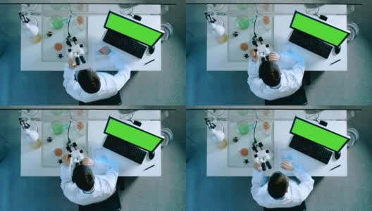 一个科学家在个人电脑上工作的俯视图，绿色的屏幕，观察显微镜和用图表把结果写在文件上。高清在线视频素材下载