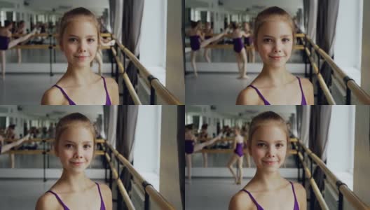 特写的美丽的小女孩在紧身衣站在芭蕾舞班，微笑着看着相机。其他学生在后台做练习。高清在线视频素材下载