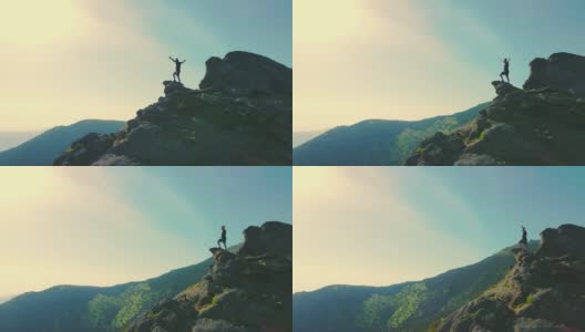Aeria绕着一个站在山顶的男人飞着，他举起双手望向远方。一个游客的剪影在岩石上看未来在日落高清在线视频素材下载