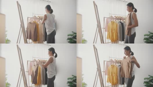 年轻的亚洲妇女在衣架上挑选衣服，在家里的客厅里穿衣照镜子。高清在线视频素材下载