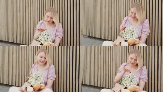 金发短发，涂着红色唇膏的时髦女人在户外放松。可爱的女孩与一束洋甘菊和纸袋法国法棍。看着镜头，开玩笑地扯下花瓣，吹着风高清在线视频素材下载