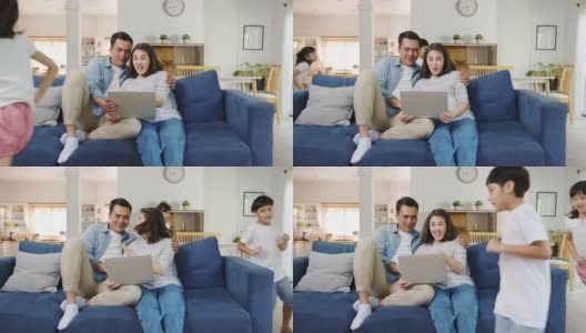 亚洲家庭爸爸和妈妈坐在沙发上享受笔记本电脑上的网上购物，而女儿和儿子在家里客厅的沙发上边玩边大喊大叫。高清在线视频素材下载