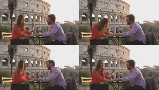 日落时分，浪漫优雅的情侣坐在罗马斗牛场前的餐厅桌前，手里拿着一杯红酒，手拉手交谈高清在线视频素材下载