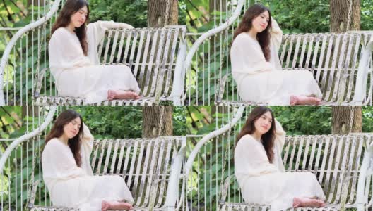 美丽的中国年轻女子穿着白色的裙子，光着脚坐在摇椅上摇摆睡觉，美丽的夏天，4k镜头，慢镜头。高清在线视频素材下载