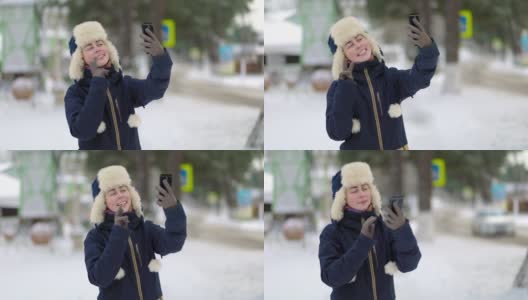 一名戴着帽子、戴着耳罩、穿着夹克、面带微笑的年轻女子的肖像，她用智能手机自拍。背景是白雪覆盖的街道高清在线视频素材下载