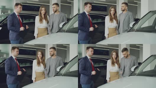 友好的汽车销售员正在与自信的年轻人告诉他的新车型，而漂亮的女人是站在她的丈夫旁边，抱着他的手臂和微笑。高清在线视频素材下载