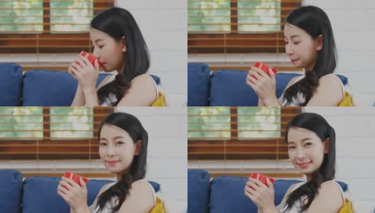 近距离的年轻亚洲女人拿着红色的咖啡杯，吸入和微笑时看着镜头，而坐在家里客厅的沙发背景，人们的饮食生活方式高清在线视频素材下载