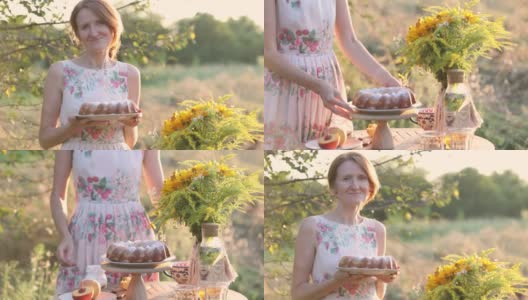 微笑的女人手拿自制的蛋糕站在餐桌旁，在傍晚的阳光照耀下的花园高清在线视频素材下载