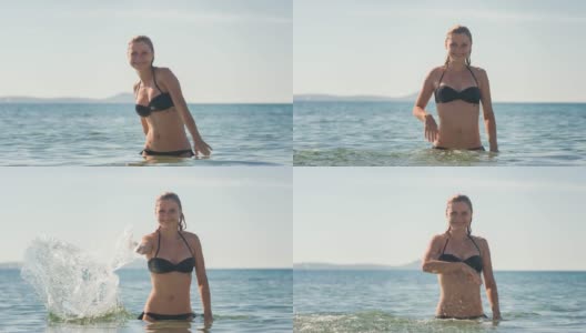 在一个炎热的阳光灿烂的日子里，一个女人在海滩上享受夏天，站在大海里，对着镜头溅起海水高清在线视频素材下载