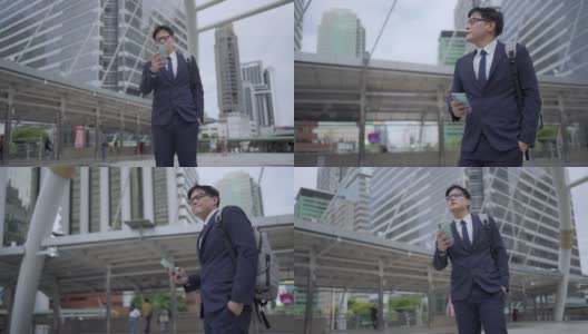 慢动作，微笑的年轻亚洲商人办公室职员穿着西装走在城市里，用智能手机发送在线信息。在市区使用手机工作或社交媒体的商人高清在线视频素材下载